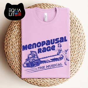 May include: A pink t-shirt with a blue graphic of a woman sitting on a chainsaw. The text "Menopausal Rage" and "The Musical" are printed on the shirt.