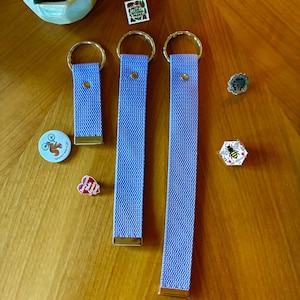 May include: Three light purple fabric wristlet keychains with gold-tone hardware, each featuring a circular ring. The set is accompanied by five small decorative pins, including designs of a squirrel, a bee, and text reading Protect Our National Parks.