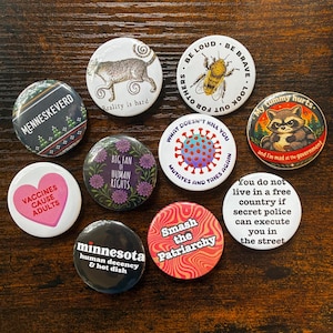 May include: A collection of round pinback buttons with various designs and text. The buttons feature illustrations of a cat, bee, raccoon, and a virus. Text includes phrases like "Reality is hard," "Big Fan of Human Rights," and "Smash the Patriarchy."