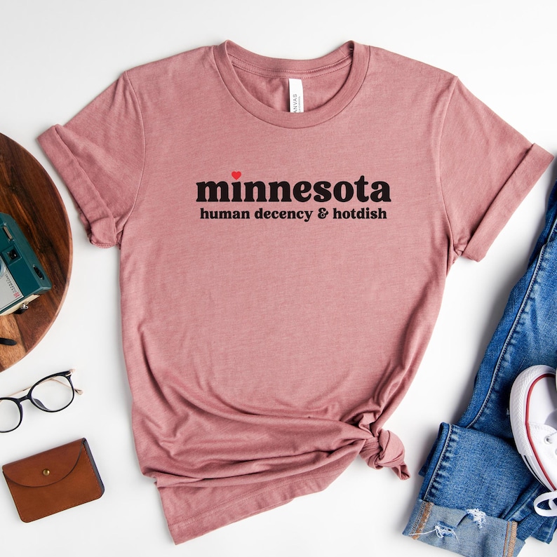 May include: A pink t-shirt with the text "minnesota human decency & hotdish" in black with a small red heart above the word "minnesota".