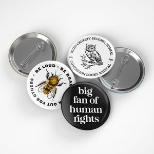 May include: A collection of five round pinback buttons. One black button reads "big fan of human rights" in white. Other buttons feature a bee, an owl, and text that reads "Be Loud, Be Brave, Out for Others" and "When Cruelty Becomes Normal, Compassion Looks Radical."