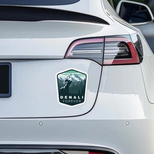 May include: A white car with a sticker on the back. The sticker is a green and blue illustration of a mountain range with a deer in the foreground. The text on the sticker reads "Denali Forever".