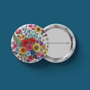 May include: Two round pinback buttons. One button features a colorful floral design with red poppies, yellow sunflowers, and bluebells. The other button is silver with a pin on the back. The background is a solid teal color.