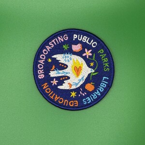 Pode incluir: Patch bordado com borda azul marinho e fundo verde. O patch apresenta as palavras "Broadcasting Public Parks Libraries Education" em um padr&atilde;o circular. Um p&aacute;ssaro branco com detalhes em laranja e amarelo est&aacute; no centro.