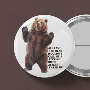 May include: A white button with a brown bear illustration. Text on the button reads "AT LEAST THE BEAR WOULDN'T CALL ME A F*CKING BITCH AFTER IT KILLED ME."