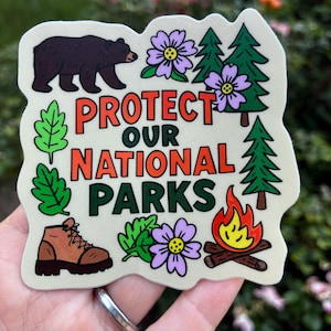 May include: A die-cut sticker featuring a brown bear, green pine trees, purple flowers, a brown hiking boot, and a campfire. The text reads PROTECT OUR NATIONAL PARKS in orange and dark green lettering against a light cream background.