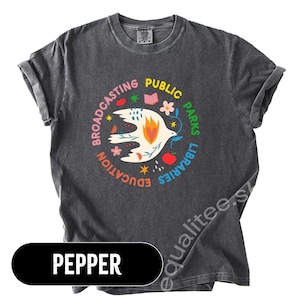 May include: Pepper-colored t-shirt with a round graphic featuring a white dove holding a branch, surrounded by colorful text: "Education, Broadcasting, Public, Parks, Libraries." The shirt has rolled-up sleeves.