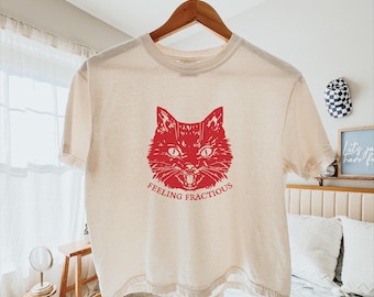 May include: A cream-colored short-sleeve t-shirt featuring a red graphic of a cat's face with its mouth open. Below the cat illustration, the text "FEELING FRACTIOUS" is printed in red capital letters. The shirt is displayed on a wooden hanger.