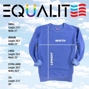 Pode incluir: Uma camisola azul periwinkle com a palavra "IGUALDADE" em letras pretas. A letra "U" da palavra &eacute; colorida com as cores do arco-&iacute;ris. As medidas da camisola s&atilde;o exibidas, com comprimento e largura em cent&iacute;metros para os tamanhos S a 2XL.