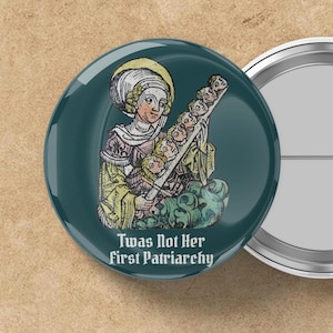 May include: A teal button with a medieval-style illustration of a woman holding a staff with several faces. The text "Twas Not Her First Patriarchy" is printed below the image. The button has a shiny, reflective surface.