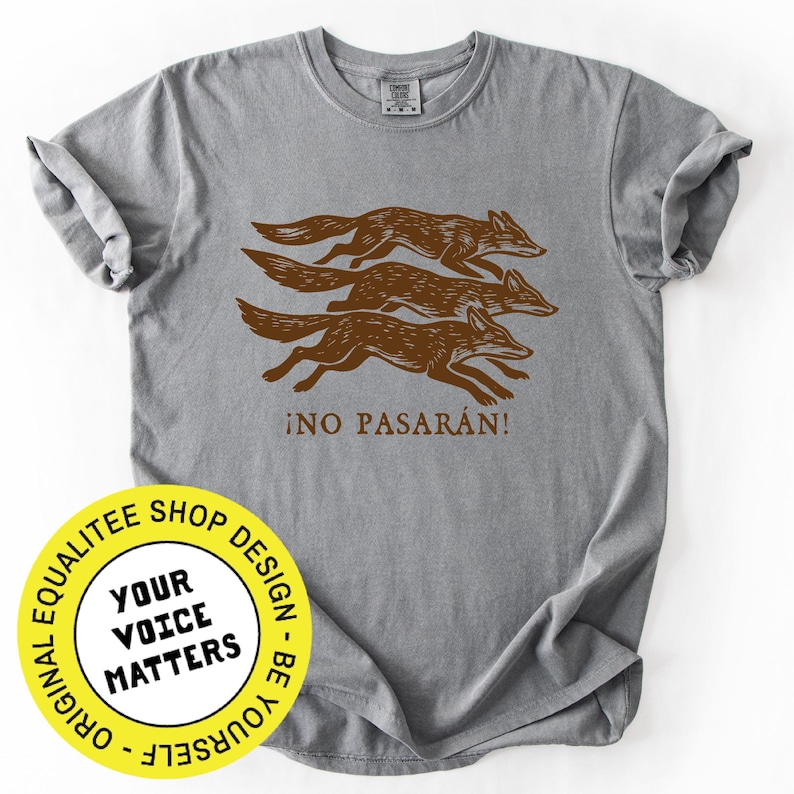 May include: A gray t-shirt featuring a brown illustration of three running foxes. The text "&iexcl;NO PASAR&Aacute;N!" is printed below the foxes. The shirt has a crew neck and short sleeves.