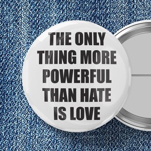May include: White button with the text "THE ONLY THING MORE POWERFUL THAN HATE IS LOVE" in black. The button is set against a blue denim background. Another button is partially visible.
