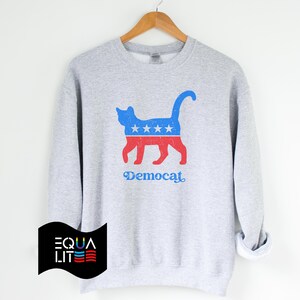 K&ouml;nnte beinhalten: Ein graues Sweatshirt mit einer blauen und roten Katzensilhouette, die ein Design aus Sternen und Streifen tr&auml;gt. Der Text "Democat" ist unter der Katze aufgedruckt.