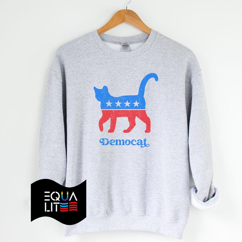K&ouml;nnte beinhalten: Ein graues Sweatshirt mit einer blauen und roten Katzensilhouette, die ein Design aus Sternen und Streifen tr&auml;gt. Der Text "Democat" ist unter der Katze aufgedruckt.