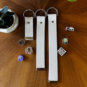 May include: Three off-white woven fabric key fobs of varying lengths, each featuring a silver-toned metal split ring at the top and a metal clamp at the bottom. Several small decorative enamel pins are scattered nearby, including designs of a bee, a bear, and text.