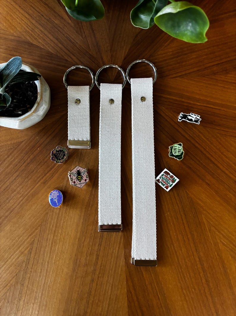 May include: Three off-white woven fabric key fobs of varying lengths, each featuring a silver-toned metal split ring at the top and a metal clamp at the bottom. Several small decorative enamel pins are scattered nearby, including designs of a bee, a bear, and text.