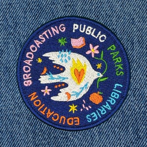 May include: Embroidered patch with a blue border and a white bird design. The patch includes the words "Broadcasting Public Education Parks Libraries" in a circular pattern. Colorful accents include a flower, book, and star.