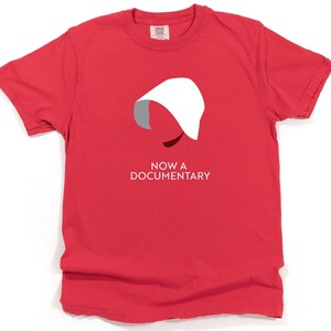 Puede incluir: Camiseta roja con una silueta blanca de una mujer con una capucha blanca y el texto "NOW A DOCUMENTARY".