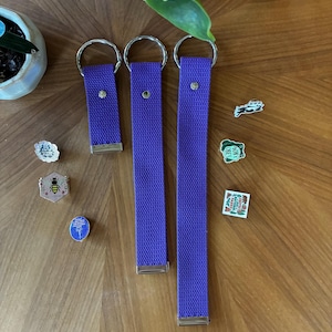 May include: Three purple fabric key fobs of varying lengths, each featuring a silver-toned metal ring at the top and a silver-toned metal clamp at the bottom. Six assorted enamel pins are arranged around the key fobs, including designs of a bee, a frog, and text.