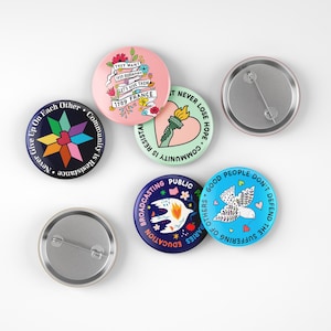 May include: A collection of colorful round buttons with various designs and text. One button is pink with floral accents and the text "They Want 1939 Germany Lets Give Them 1789 France". Other buttons have a torch, a dove, and geometric shapes.