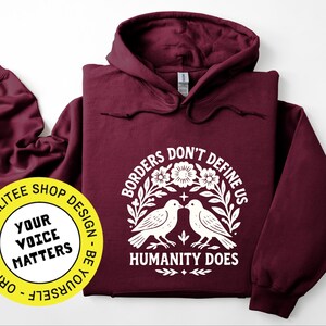 Pode incluir: Um moletom vinho com um gr&aacute;fico branco que diz "Borders Don't Define Us, Humanity Does" com dois p&aacute;ssaros e elementos florais. Um adesivo amarelo com o texto "Your Voice Matters" &eacute; vis&iacute;vel.