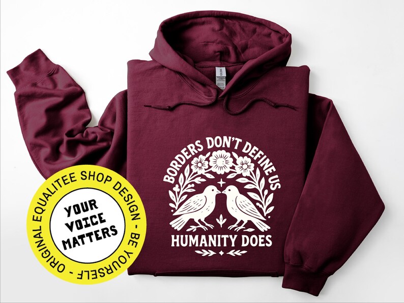 Pode incluir: Um moletom vinho com um gr&aacute;fico branco que diz "Borders Don't Define Us, Humanity Does" com dois p&aacute;ssaros e elementos florais. Um adesivo amarelo com o texto "Your Voice Matters" &eacute; vis&iacute;vel.
