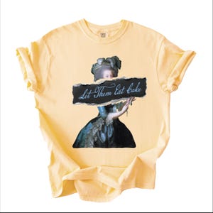 Pu&ograve; includere: T-shirt giallo chiaro con un'illustrazione vintage di una donna che tiene in mano un cartello con la scritta "Let Them Eat Cake". La donna indossa un abito blu e nero. La maglietta ha maniche corte e girocollo.