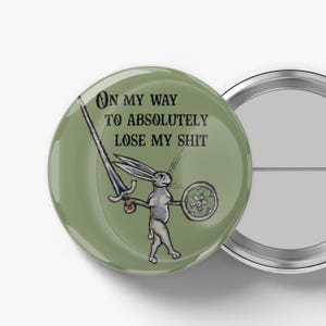 May include: A round, green button with the text "ON MY WAY TO ABSOLUTELY LOSE MY SHIT." The button features a medieval-style illustration of a rabbit holding a sword and shield. The button is next to a silver-colored button.