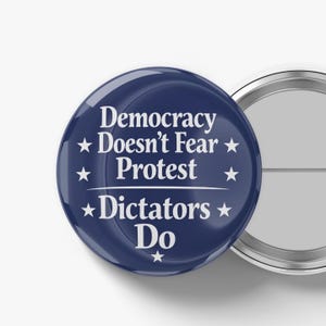 May include: A navy blue button with white text that reads "Democracy Doesn't Fear Protest Dictators Do." The button has a shiny finish and a silver pin back. The design includes white star accents.