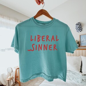 May include: A turquoise t-shirt with the words "LIBERAL SINNER" in red lettering. The shirt is hanging on a wooden hanger. The background includes a bed, a small table, and decorative items.