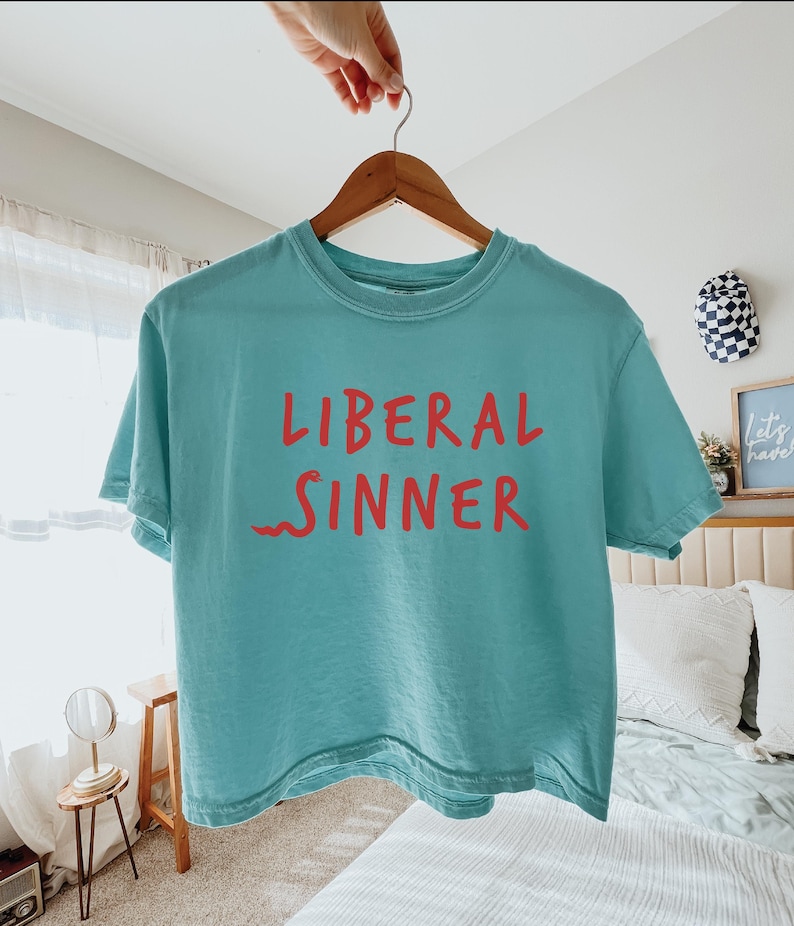 May include: A turquoise t-shirt with the words "LIBERAL SINNER" in red lettering. The shirt is hanging on a wooden hanger. The background includes a bed, a small table, and decorative items.