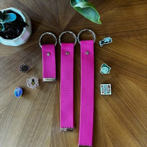 May include: Three bright pink fabric wristlet keychains of varying lengths, each featuring a silver metal split ring and a silver metal end clamp. Beside them are several small enamel pins, including a bee, a frog, and a national park emblem.