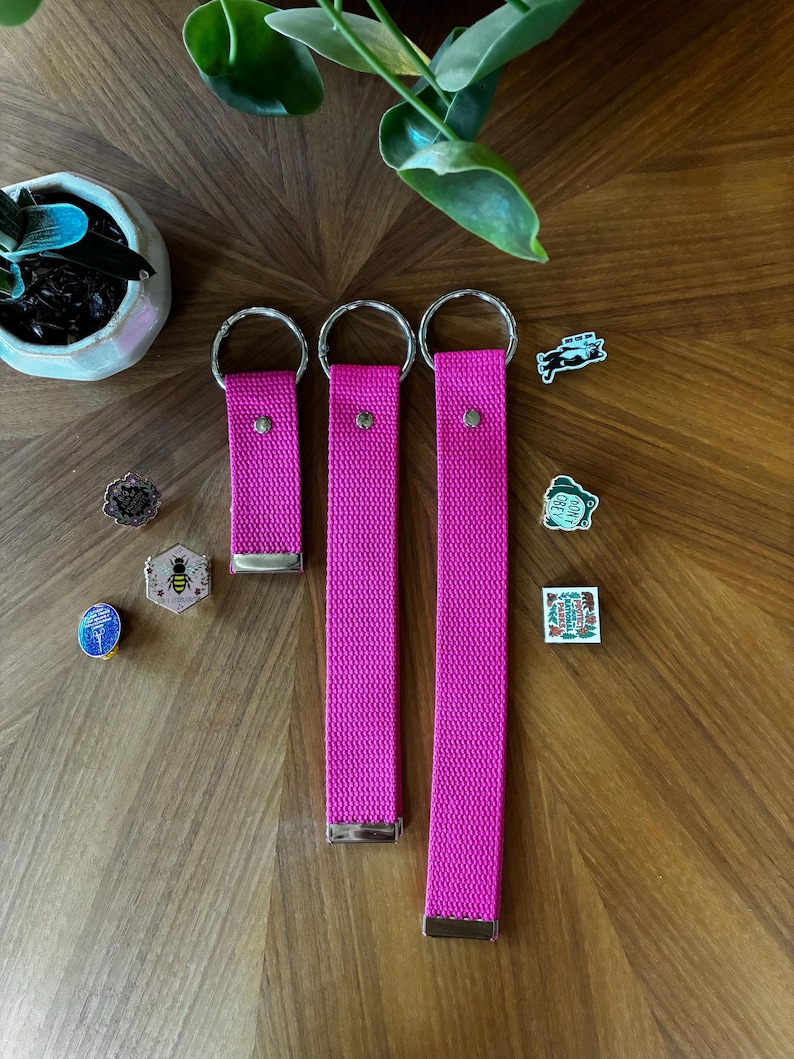 May include: Three bright pink fabric wristlet keychains of varying lengths, each featuring a silver metal split ring and a silver metal end clamp. Beside them are several small enamel pins, including a bee, a frog, and a national park emblem.