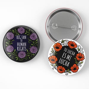 May include: Three round buttons with graphic designs. One button has the text "BIG FAN OF HUMAN RIGHTS" surrounded by purple flowers and green vines. Another button has the text "TU LUCHA ES MI LUCHA" surrounded by orange flowers and green vines.