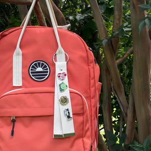 May include: A coral-colored backpack with cream-colored straps and a circular black and white sun logo patch. A cream-colored fabric keychain hangs from the strap, featuring four enamel pins: a red heart, a green frog, a gold circle, and a cat.