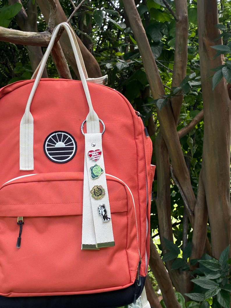 May include: A coral-colored backpack with cream-colored straps and a circular black and white sun logo patch. A cream-colored fabric keychain hangs from the strap, featuring four enamel pins: a red heart, a green frog, a gold circle, and a cat.