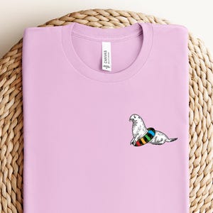 May include: A light purple t-shirt with a black and white illustration of a seal wearing a rainbow inner tube.