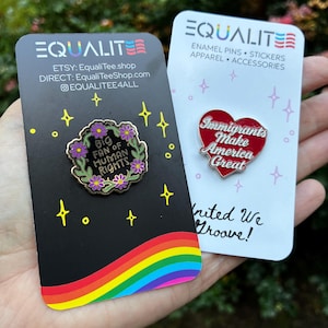 Pu&ograve; includere: Due spille smaltate su cartoncini. Una spilla recita "Big Fan of Human Rights" circondata da fiori. L'altra spilla &egrave; a forma di cuore e recita "Immigrants Make America Great". I cartoncini hanno disegni arcobaleno e stelle.