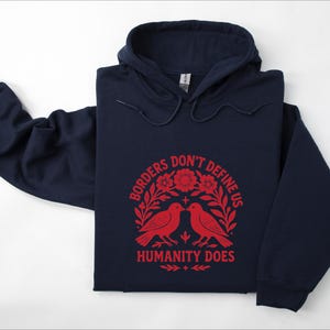 Pode incluir: Camisola com capuz azul marinho com o texto vermelho "Borders Don't Define Us Humanity Does". O design inclui dois p&aacute;ssaros vermelhos e elementos florais. A camisola tem capuz com cord&atilde;o e mangas compridas. Medidas em cm.
