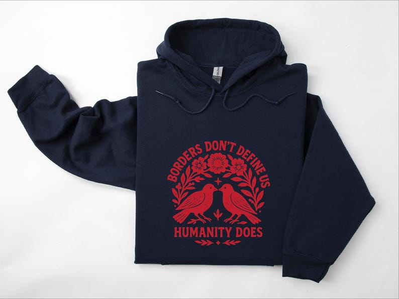 Pode incluir: Camisola com capuz azul marinho com o texto vermelho "Borders Don't Define Us Humanity Does". O design inclui dois p&aacute;ssaros vermelhos e elementos florais. A camisola tem capuz com cord&atilde;o e mangas compridas. Medidas em cm.