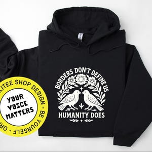 Pode incluir: Moletom preto com um gr&aacute;fico branco. O gr&aacute;fico apresenta dois p&aacute;ssaros voltados um para o outro, cercados por flores e o texto "Borders Don't Define Us, Humanity Does". Um adesivo amarelo com texto est&aacute; no canto inferior esquerdo.