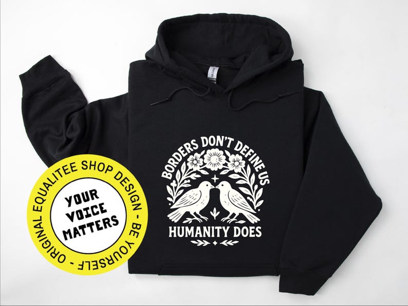 Pode incluir: Moletom preto com um gr&aacute;fico branco. O gr&aacute;fico apresenta dois p&aacute;ssaros voltados um para o outro, cercados por flores e o texto "Borders Don't Define Us, Humanity Does". Um adesivo amarelo com texto est&aacute; no canto inferior esquerdo.