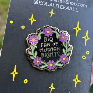 Pu&ograve; includere: Spilla smaltata nera con bordi dorati, con la scritta "Big Fan of Human Rights", circondata da una ghirlanda di foglie verdi e fiori viola. Su sfondo nero con dettagli di stelle e punti gialli.