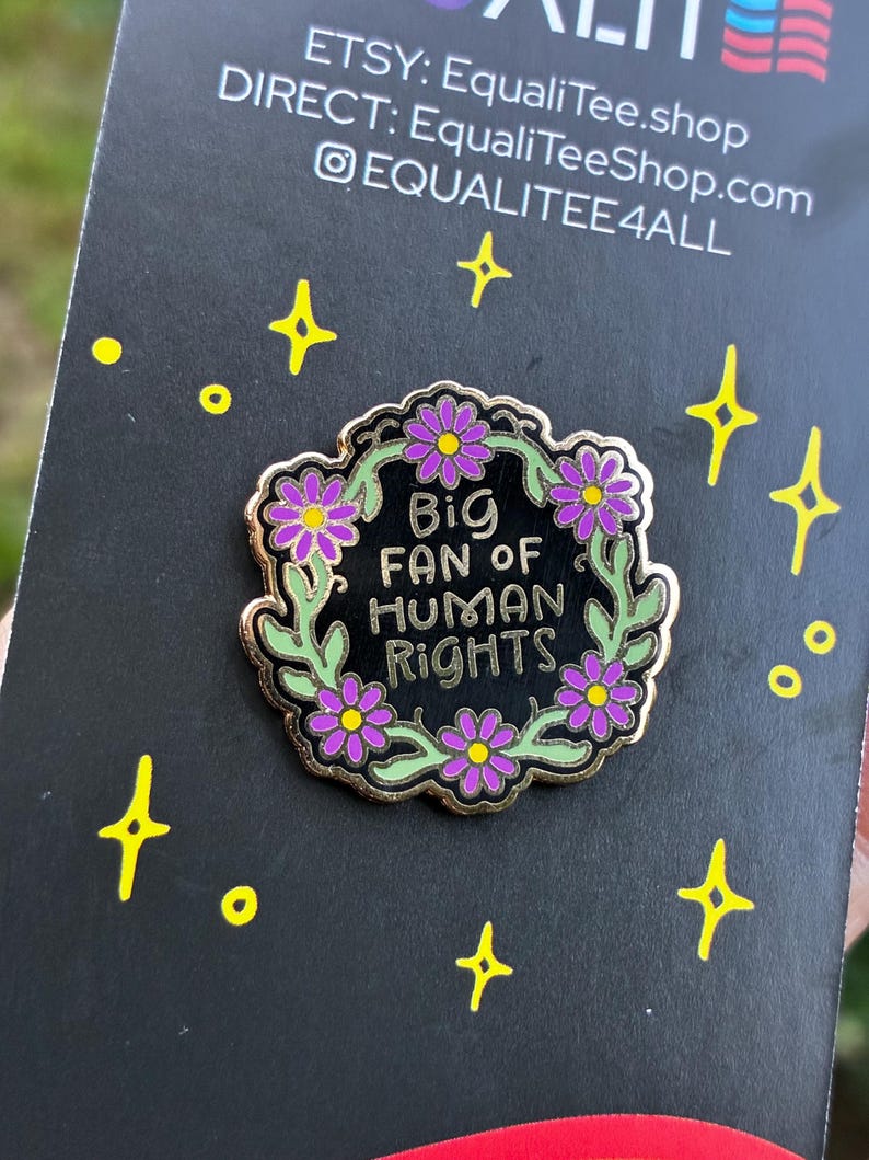 Pu&ograve; includere: Spilla smaltata nera con bordi dorati, con la scritta "Big Fan of Human Rights", circondata da una ghirlanda di foglie verdi e fiori viola. Su sfondo nero con dettagli di stelle e punti gialli.