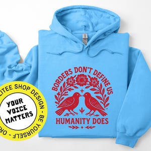 Pode incluir: Um moletom azul claro com capuz e mangas compridas. A frente apresenta um gr&aacute;fico vermelho com dois p&aacute;ssaros, flores e o texto "BORDERS DON'T DEFINE US HUMANITY DOES". Um adesivo amarelo com texto est&aacute; no canto inferior esquerdo.