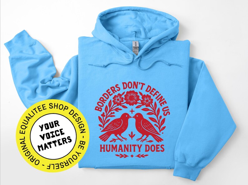 Pode incluir: Um moletom azul claro com capuz e mangas compridas. A frente apresenta um gr&aacute;fico vermelho com dois p&aacute;ssaros, flores e o texto "BORDERS DON'T DEFINE US HUMANITY DOES". Um adesivo amarelo com texto est&aacute; no canto inferior esquerdo.