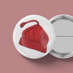May include: A red knit hat illustration on a white button. The hat has a folded brim and a braided tassel. The button is round and has a shiny metal edge. The background is a solid, muted pink.