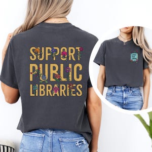 May include: A dark gray t-shirt with the text "SUPPORT PUBLIC LIBRARIES" in yellow with colorful floral accents. The back of the shirt features the same text in a smaller size with a floral border.