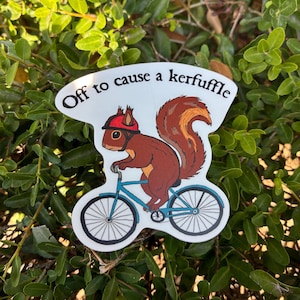 May include: A die-cut sticker featuring a brown squirrel wearing a red helmet while riding a blue bicycle. The text "Off to cause a kerfuffle" is printed in black letters along the top curve of the white sticker background.