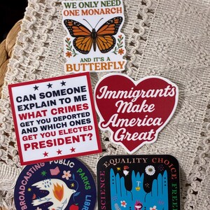 Op de afbeelding: Een verzameling kleurrijke stickers met diverse ontwerpen en tekst. E&eacute;n sticker toont een monarchvlinder, een andere een hart met de tekst "Immigrants Make America Great". Andere stickers bevatten teksten over politiek en sociale kwesties.